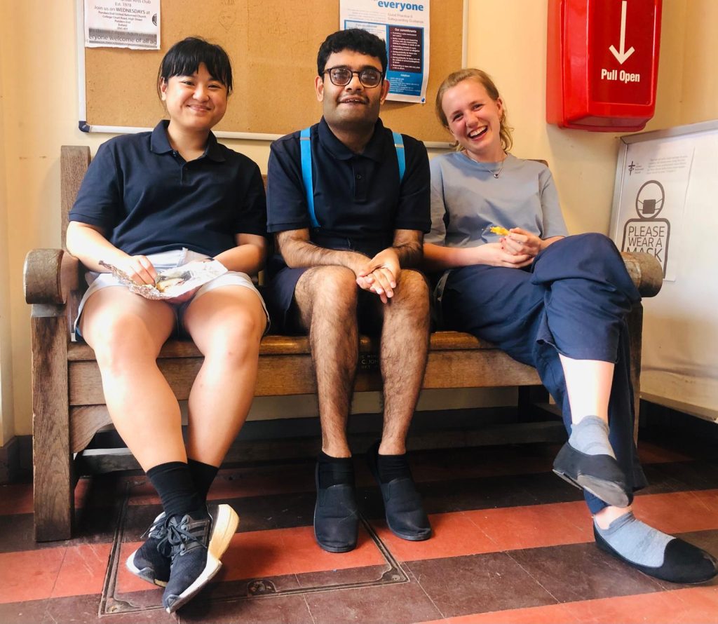 Millie sat with some of the costumed cast of Quixote. She is sat on the far right of the image and wearing her costume of a light blue t-shirt, and navy trousers. Everyone is smiling and some are finishing off a snack before going to perform the first of our shows. 